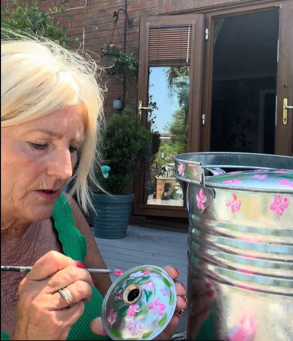 Sally painting pink blossom on the lid of a galvanised tub, outdoors on her decking in the sun
